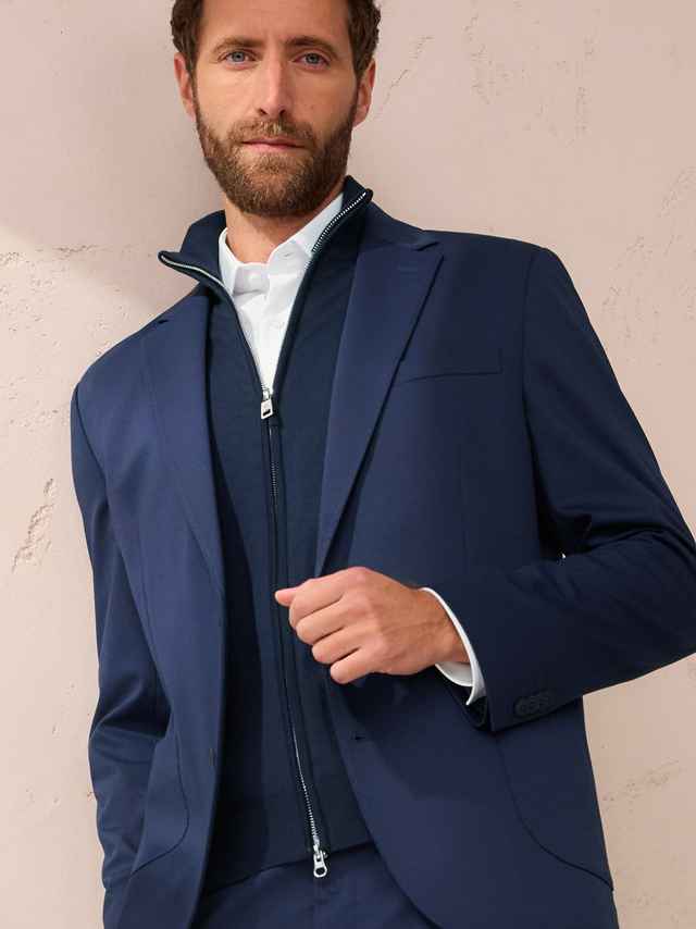 Mann mit Bart in blauem Anzug, darunter ein dunkelblauer Pullover mit Reißverschluss, vor einer hellen Wand stehend.