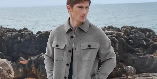 Mann in grauer Jacke steht vor felsiger Küste mit Blick aufs Meer.