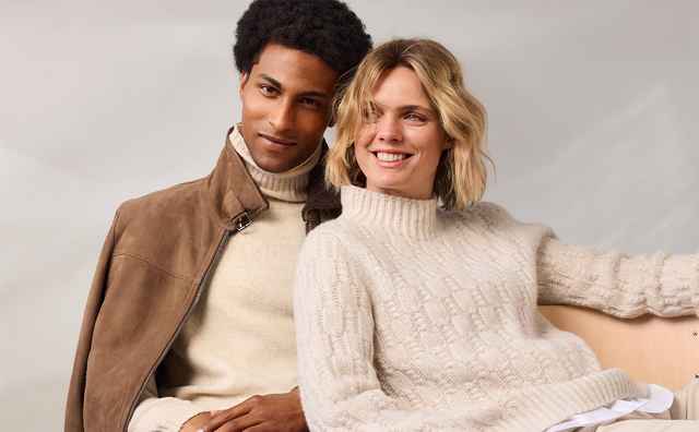 Ein Mann und eine Frau sitzen lächelnd nebeneinander auf einem Sofa, beide tragen Creme-Pullover.