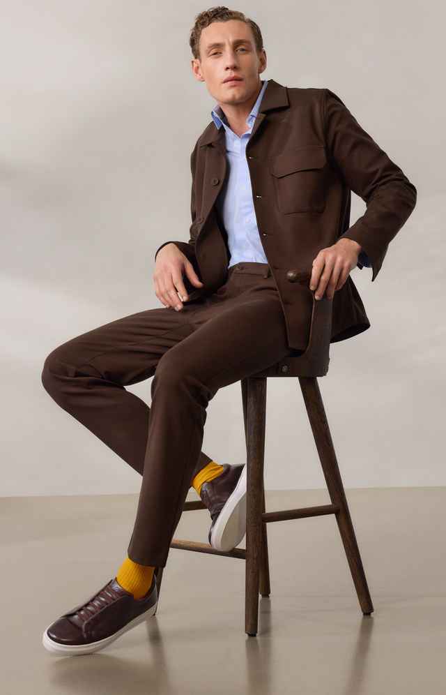 Ein Mann in einem braunen Anzug mit hellblauem Hemd sitzt auf einem Stuhl. Er trägt gelbe Socken und braune Slipper.