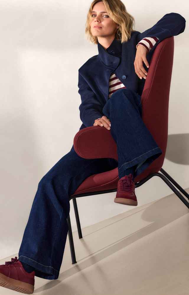 Frau in dunkelblauer Jacke und Jeans sitzt lässig in rotem Sessel. Sie trägt gestreiftes Oberteil und rote Sneaker.