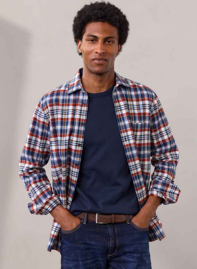 Mann mit Afro trägt kariertes Hemd über dunkelblauem T-Shirt und Jeans, die Hände in den Hosentaschen, vor grauem Hintergrund.