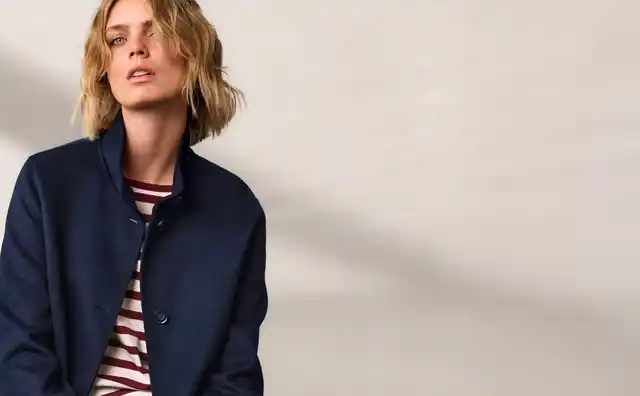 Person mit blondem Haar trägt ein gestreiftes Shirt und eine dunkelblaue Jacke, sitzt vor einer hellen Wand.