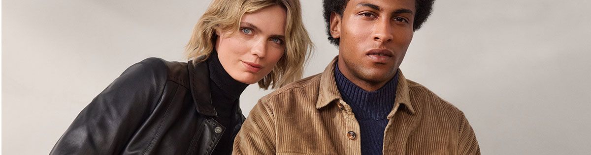 Zwei Personen in herbstlicher Kleidung stehen nebeneinander, eine Frau in einer Lederjacke und ein Mann in einer braunen Cordjacke und einem blauen Pullover.