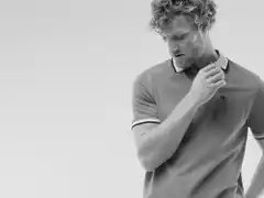 Mann mit lockigem Haar trägt ein Poloshirt und blickt nach unten. Schwarz-Weiß-Fotografie mit schlichtem Hintergrund.