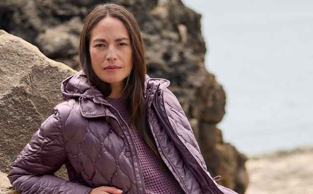 Frau in violetter Steppjacke und Strickpullover lehnt an einem Felsen, trägt graue Jeans und braune Stiefeletten, im Freien.