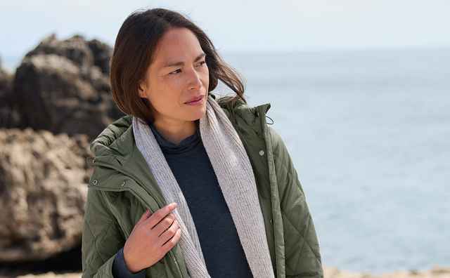 Frau in grüner Steppjacke und Jeans steht entspannt auf einem Felsen am Meer; sie trägt einen grauen Schal, der lose um den Hals hängt.