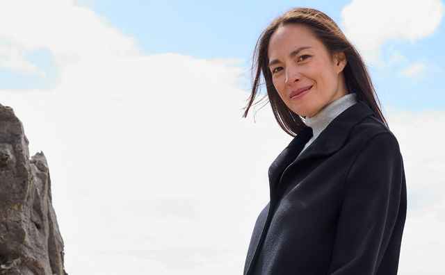 Frau in schwarzem Mantel und blauer Jeans lächelt am felsigen Strand mit Meer im Hintergrund.