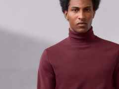 Mann mit lockigem Haar trägt einen bordeauxroten Rollkragenpullover vor grauem Hintergrund.
