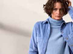 Person mit kurzem, lockigem Haar trägt einen blauen Rollkragenpullover und ein kariertes Hemd.
