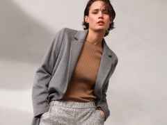 Frau in grauem Blazer und braunem Pullover, mit kurzen Haaren, schaut in die Kamera. Hintergrund neutral grau.