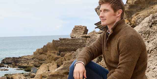 Ein Mann in einem braunen Pullover sitzt auf Felsen am Meer und blickt in die Ferne. Der Himmel ist leicht bewölkt.