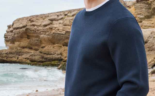 Mann in dunkelblauem Pullover steht an einem felsigen Strand vor dem Meer.
