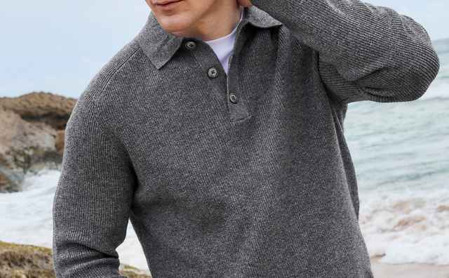Ein Mann in grauem Pullover steht am felsigen Strand, schaut nachdenklich zur Seite.