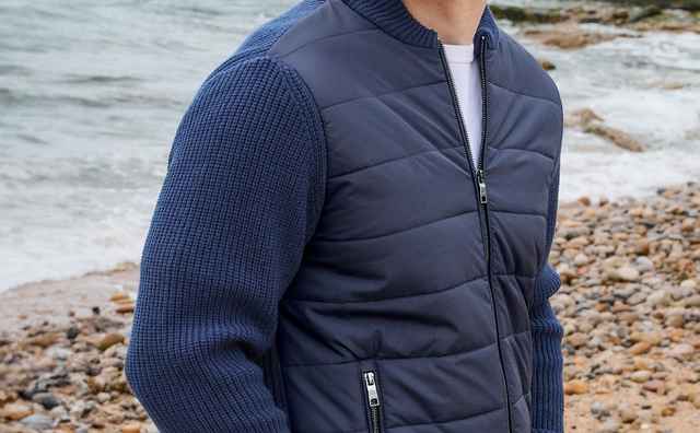Mann in blauer Jacke und Jeans steht am steinigen Strand, blickt nach links. Im Hintergrund sind Meer und Felsen zu sehen.