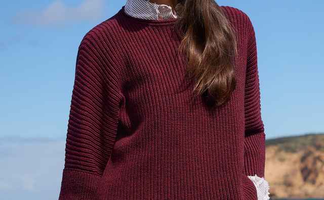 Frau am Meer trägt einen weinroten Pullover und eine gestreifte Bluse. Sie blickt lächelnd in die Ferne, der Himmel ist klar und blau.