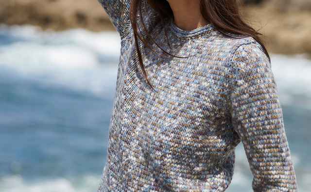 Frau in Strickpullover am Meer, braunes Haar, Hand auf Kopf, Blick in die Ferne, Felsen im Hintergrund.