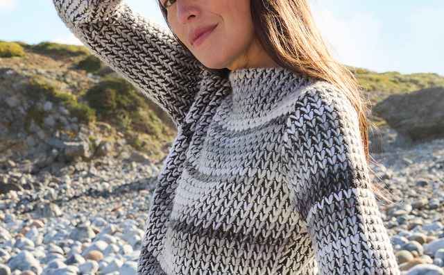 Frau am Steinstrand in gestricktem Pullover, hebt den Arm gegen die Sonne, um ihr Gesicht zu schützen. Blau himmel im hintergrund.