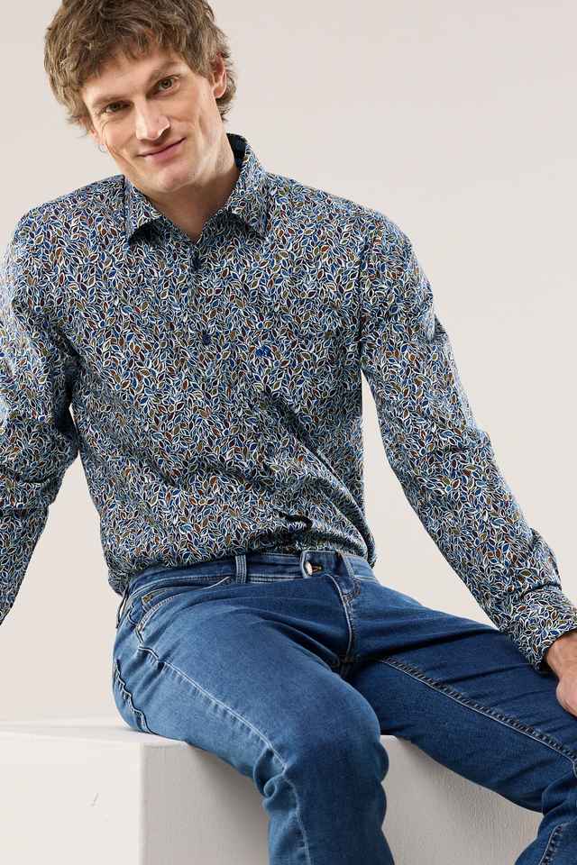 Mann in einem gemusterten Hemd und blauer Jeans sitzt auf einer weißen Kiste und lächelt in die Kamera.