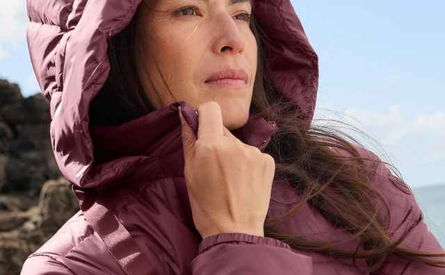 Frau in einer merlotfarbenen, wattierten Outdoorjacke mit großer Kapuze und sportlichem Stil.