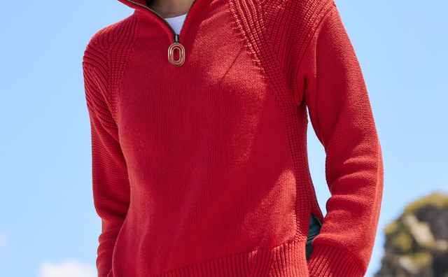 Frau in rotem Pullover mit Reißverschluss und blauer Jeans steht draußen vor einer felsigen Landschaft und blauem Himmel.