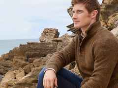 Ein Mann in einem braunen Pullover sitzt auf Felsen am Meer und blickt nachdenklich in die Ferne.