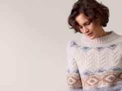 Frau mit braunem, lockigem Haar trägt einen gestrickten, gemusterten Pullover in warmen Tönen und schaut nach unten.