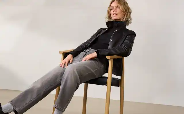 Eine Frau in schwarzer Lederjacke und grauen Jeans sitzt lässig auf einem Stuhl vor einem neutralen Hintergrund.