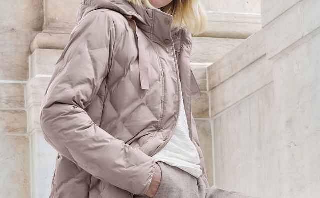 Person in einer taupefarbenen Steppjacke mit Kapuze, kombiniert mit einem weißen Oberteil und grauer Hose. Der Stil wirkt lässig und herbstlich.
