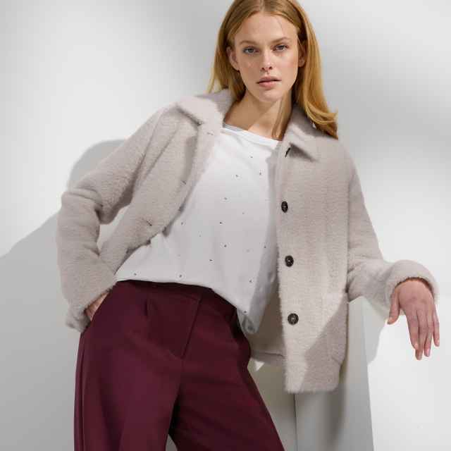 Person in einer hellen, flauschigen Jacke über einem weißen, verzierten Oberteil und einer bordeauxroten Hose. Der Stil wirkt elegant und winterlich.