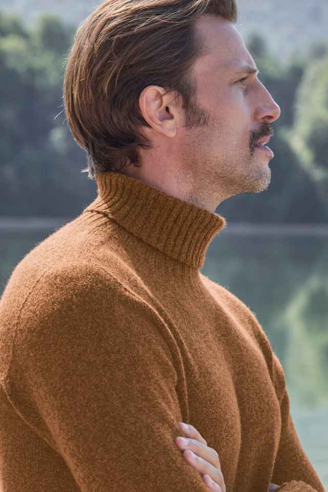 Seitenprofil eines Mannes mit braunem Rollkragenpullover, der vor einem See und Wald im Hintergrund steht.