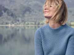 Frau mit blonden Haaren in einem blauen Pullover sitzt lächelnd vor einem See mit bergiger Landschaft im Hintergrund.