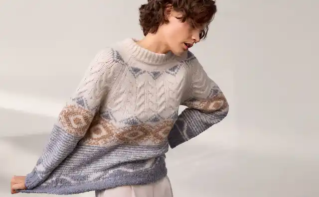 Person mit lockigem Haar trägt einen gestrickten Pullover mit geometrischem Muster in Creme, Blau und Braun vor einem neutralen Hintergrund.