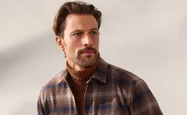 Mann mit braunem Rollkragenpullover und karierter Hemdjacke, den Blick nach links gewandt, steht vor einem neutralen Hintergrund.