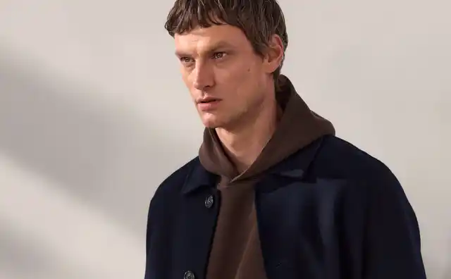 Ein Mann trägt einen dunkelblauen Mantel über einem braunen Hoodie und brauner Hose, steht vor einem grauen Hintergrund und blickt zur Seite.