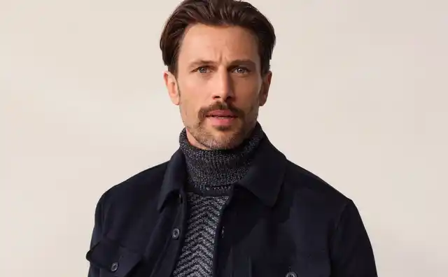 Mann in dunkelblauer Jacke und grauem Rollkragenpullover, steht lässig vor hellem Hintergrund.