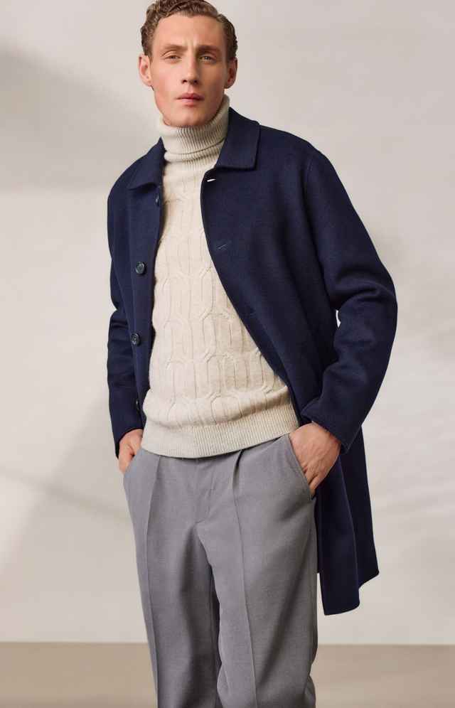 Mann trägt blauen Mantel, cremefarbenen Rollkragenpullover und graue Hose. Hände in den Taschen, steht vor hellem Hintergrund.