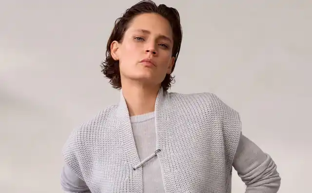 Frau mit kurzem dunklem Haar trägt einen grauen Strickpullover über einem hellen Shirt und blaue Jeans mit Knöpfen, vor neutralem Hintergrund.