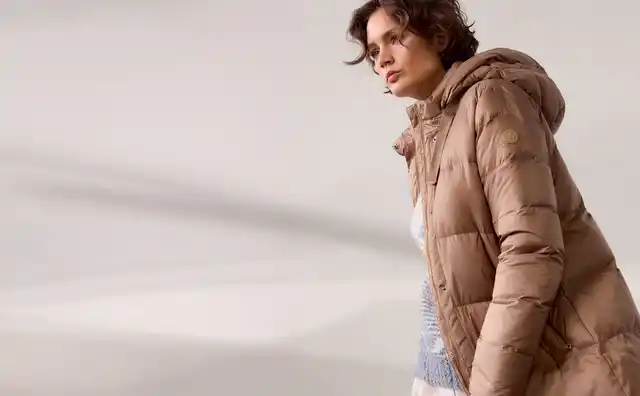 Frau in beigem Wintermantel steht in einem hellen Raum. Sie trägt einen grauen Pullover und weiße Hose, schaut zur Seite.