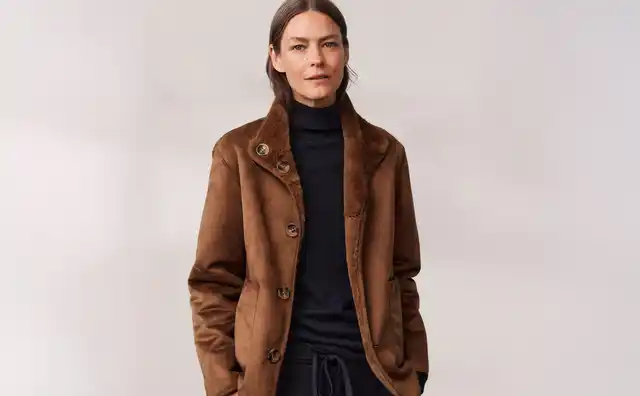 Eine Frau trägt eine braune Jacke und schwarzen Rollkragenpullover, mit den Händen in den Taschen vor einem neutralen Hintergrund.