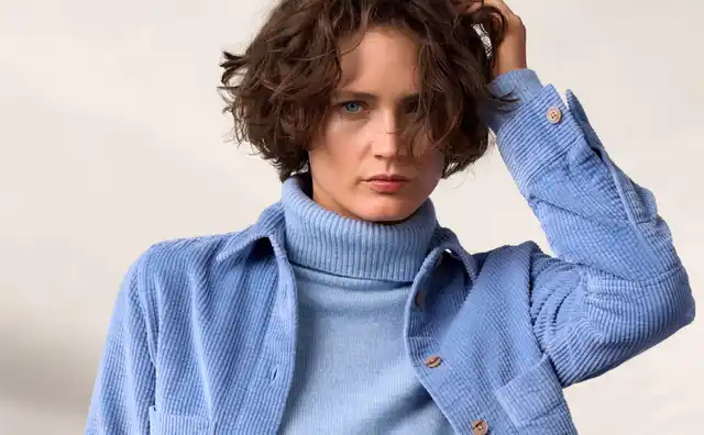 Eine Frau trägt einen blauen Rollkragenpullover, ein blaues Oberhemd und eine dunkelblaue Hose, steht vor einem schlichten Hintergrund.