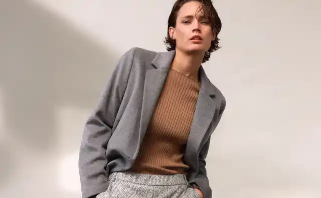 Frau in grauem Blazer und braunem Pullover, mit hellen gemusterten Hosen, posiert vor einem neutralen Hintergrund.