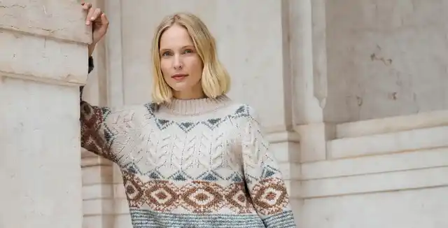 Frau mit blondem Haar trägt einen gemusterten Strickpullover und lehnt an einer Steinsäule.