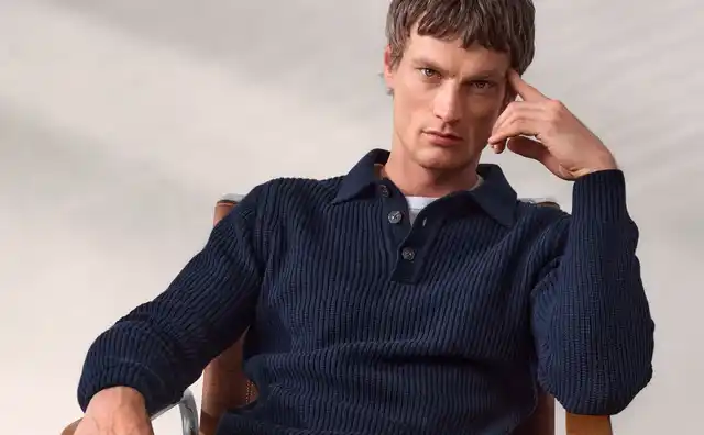 Ein Mann sitzt auf einem Stuhl, trägt einen dunkelblauen Pullover, beige Hose und weiße Turnschuhe.