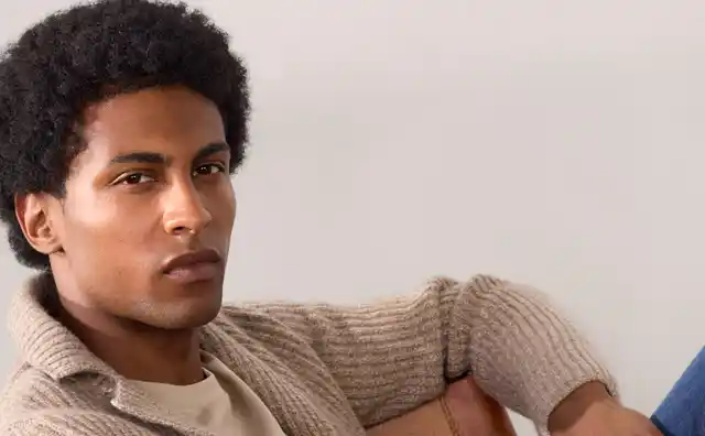 Ein Mann in einem beigen Strickpullover lehnt entspannt auf einem Stuhl, trägt ein T-Shirt und Jeans.