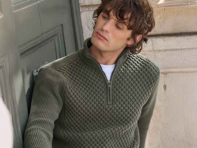 Ein Mann in einem olivgrünen Strickpullover lehnt an einer grünen Tür und schaut zur Seite.
