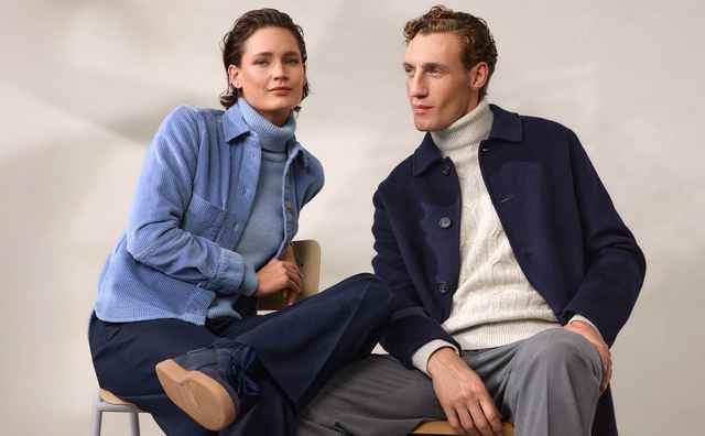 Zwei Personen in stilvoller Herbstkleidung sitzen nebeneinander. Eine trägt eine blaue Jacke, die andere einen dunklen Mantel mit Rollkragenpullover.